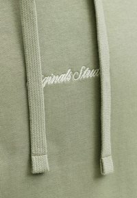 Olivi vihreä huppari, jossa on teksturoitu kangas, varustettuna nyörillä ja valkoisella brodeeratulla tekstillä, jossa lukee "Originals Style." Yksinkertainen muotoilu, ilman kuvioita.