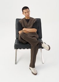 Brun strikket vest over en svart T-skjorte, kombinert med brune bukser og beige sneakers. Sittende på en svart skinnstol med kromben.
