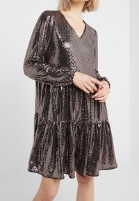 Robe pailletée avec un décolleté en V, des manches longues et une jupe à volants. Présente un motif métallique rose doré et une texture douce.