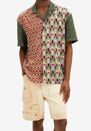 Homme portant une chemise à manches courtes à boutons avec des motifs géométriques et ovales mélangés, associée à un short cargo beige avec plusieurs poches.