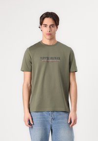 T-shirt de algodão verde-oliva com decote redondo e mangas curtas, com o logótipo "TOMMY HILFIGER" impresso em cores contrastantes na frente.