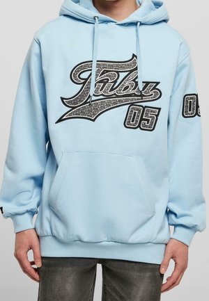 Persona che indossa una felpa con cappuccio azzurro chiaro con un grande logo "FUBU 05" nero e grigio sul davanti e una toppa abbinata sulla manica sinistra.