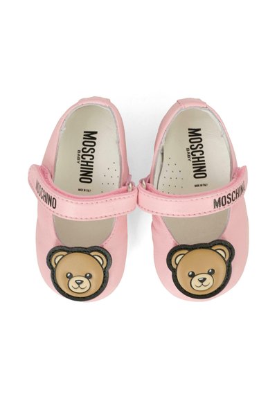De petits chaussons en cuir rose clair pour bébé avec une sangle Velcro, arborant un emblème de tête d'ours rond sur l'embout et une marque "Moschino" sur la sangle.