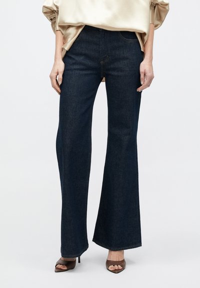 Jeansi evazați de culoare albastru închis, din denim, cu un design clasic cu cinci buzunare. Purtați cu o bluză bej deschis și tocuri maro decupate la vârf.