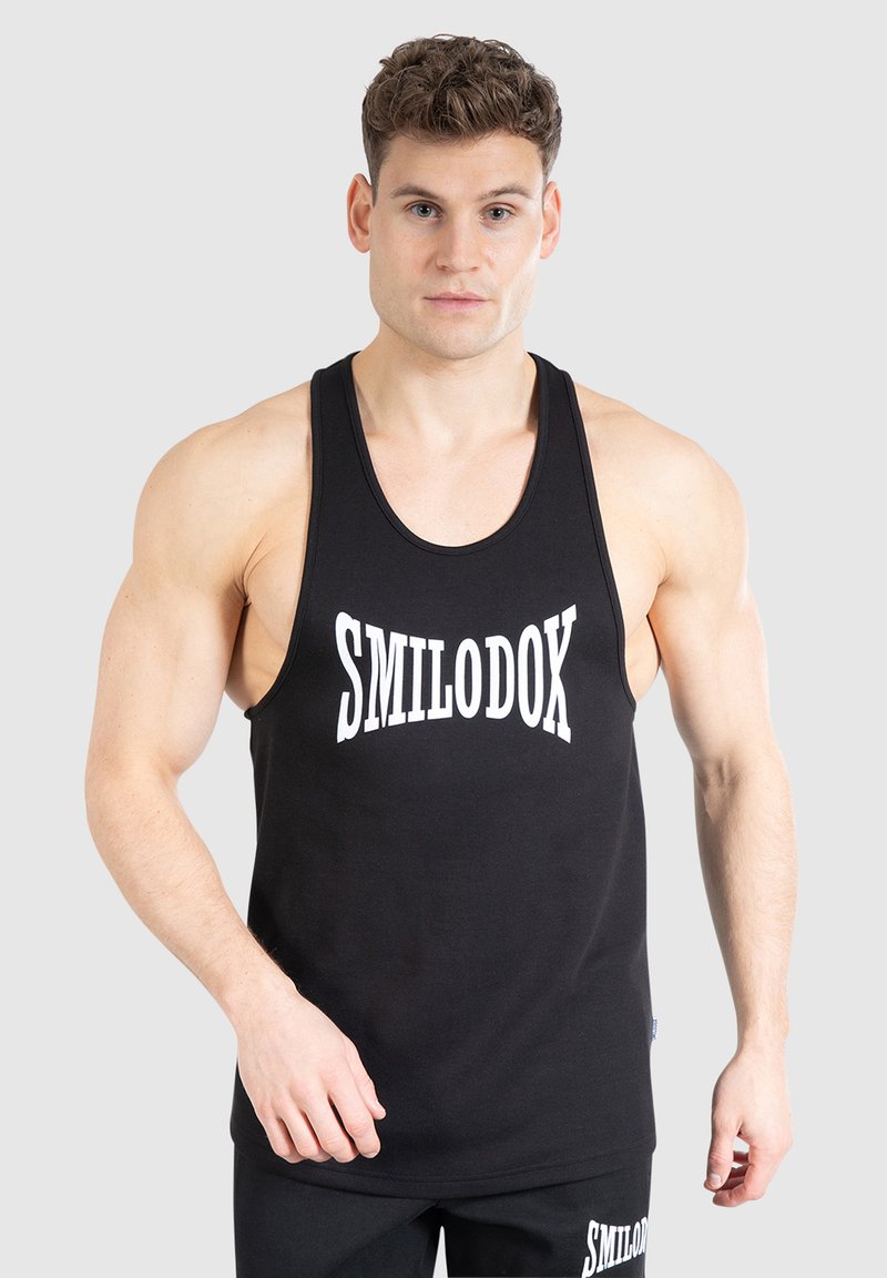 Spierlijke man in een zwart mouwloos sportshirt en shorts met "SMILODOX" in het wit erop gedrukt, staand tegen een effen achtergrond.