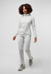 Lichtgrijze sportoutfit met een ritsjack met hoge kraag en taps toelopende broek. Het materiaal lijkt lichtgewicht en ademend.