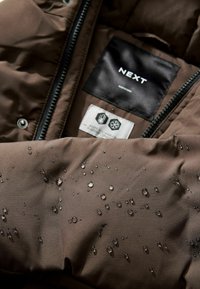 Interno di giacca puffer marrone scuro con una texture liscia. Presenta una superficie idrorepellente con gocce d'acqua visibili e un'etichetta nera.