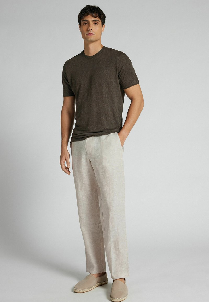 Mann trägt dunkelbraunes kurzärmeliges T-Shirt, hellbeige Leinenhose und beige Slipper, steht vor einem einfarbigen grauen Hintergrund.