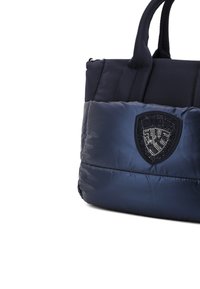Bolso tote acolchado en color marino con superficie texturizada y acolchada; presenta un logo negro en forma de escudo con adornos decorativos y asas robustas.