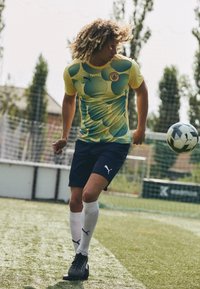 Žlutý a tyrkysový vzorovaný fotbalový dres, námořnické šortky, bílé ponožky s černým logem, na hráči, který žongluje s fotbalovým míčem na trávě.