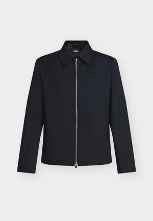 Veste noire à manches longues avec col et fermeture éclair argentée à l'avant, étiquette de la marque visible à l'intérieur du col, posée à plat sur fond blanc.