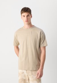 T-shirt en coton beige à manches courtes, avec un graphique centré d'un phare et du texte. Le modèle porte un pantalon camouflé clair.