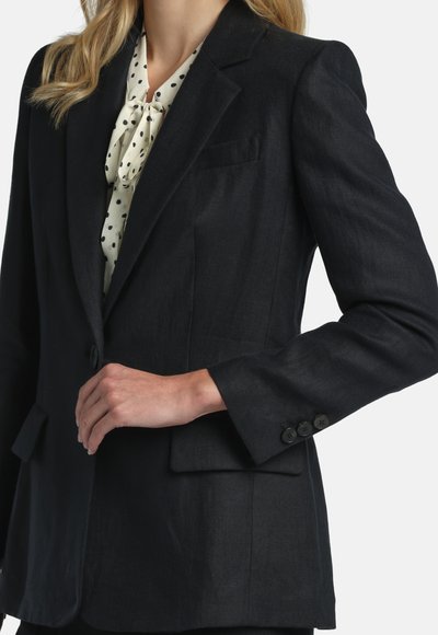 Woman wearing a black tailored blazer over a white blouse with black polka dots, with her hand resting on her waist.