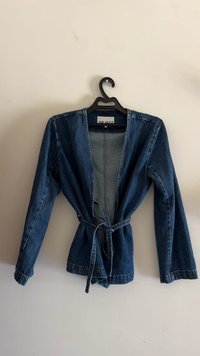 Chaqueta de denim de color azul oscuro con mangas largas, abertura frontal y cinturón de lazo en la cintura. Presenta un dobladillo sin tratar y un detalle de costura mínima.