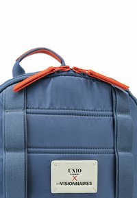 Mochila azul con textura acolchada, tiradores de cremallera naranjas y una etiqueta blanca con el diseño "UNIO HAMBURG X LES VISIONNAIRES".