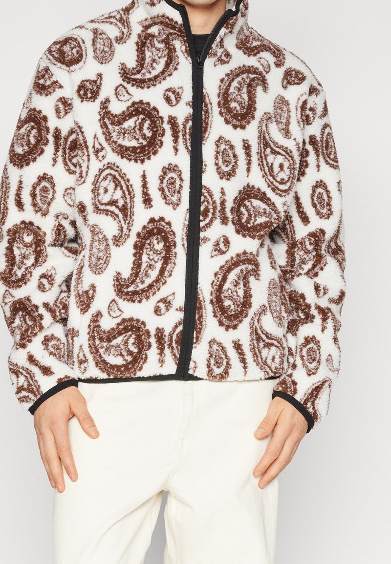 Fleece bunda s bílým základem a hnědým paisley vzorem, zapínání na zip a černým lemem kolem límce a manžet.