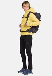 Gelbe Steppjacke mit Kapuze, schwarzer Rucksack, schwarze Hose und blaue Schuhe. Merkmale sind Reißverschlüsse, Schnallen und ein strukturiertes Gewebe.