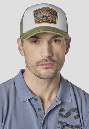 Hombre con barba clara que lleva una gorra tipo trucker blanca y oliva con parche de tigre, y una camisa polo azul clara con un gráfico negro en el pecho.