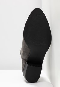 Vue de la semelle d'une botte cheville en daim gris foncé avec un bout pointu et un talon bloc empilé sur un fond blanc.