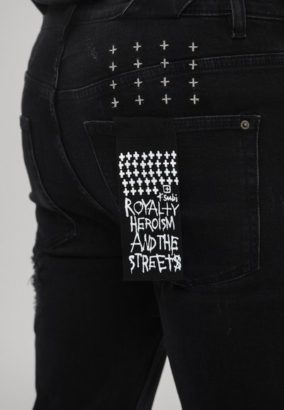 Los jeans de mezclilla negra presentan acentos de puntada cruzada en el bolsillo trasero, con una etiqueta que muestra texto gráfico sobre un patrón en blanco y negro.
