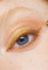Nærbilde av et øye med gulløyenskygge, som har en jevn tekstur og en definert vinget eyeliner-form, rammet inn av lyse vipper.
