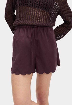 Kvinde iført mørkelilla shorts med høj talje og bølgekantede kanter samt en sort løstsiddende strikket sweater, stående med hænderne bag ryggen.