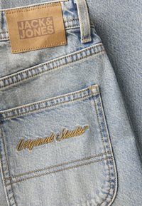 Τζιν σε ανοιχτό μπλε χρώμα με δερμάτινη ετικέτα που αναγράφει "JACK & JONES." Η πίσω τσέπη έχει το "Originals Studio" σε χρυσή ραφή.