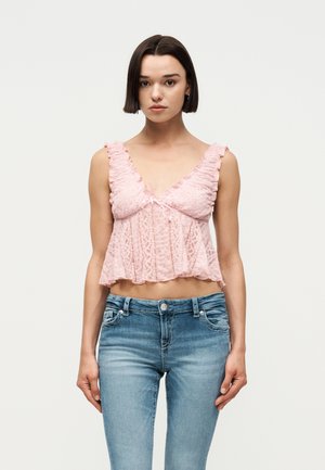 Νεαρή γυναίκα με κοντά καστανά μαλλιά που φοράει ροζ δαντελένιο αμάνικο τοπ και μπλε τζιν, στέκεται μπροστά σε απλό φωτεινό φόντο.