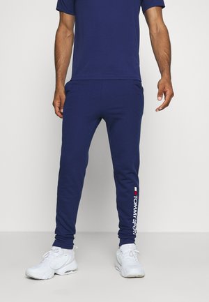 Mężczyzna ubrany w granatowe spodnie dresowe z białym i czerwonym logo „Tommy Sport” na lewej nodze oraz białe trampki, stojący na jednolitym tle.