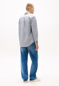 Bărbat în picioare, cu fața spre spate, purtând o cămașă cu mâneci lungi gri deschis, blugi albaștri și adidași albi pe un fundal alb simplu.