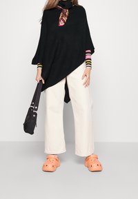 Černý pletený pončo s rolákem, barevné pruhované rukávy, v kombinaci s bílými širokými kalhotami a oranžovými dřeváky. Drží černou tašku.