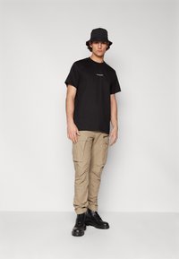Schwarzes Baumwoll-T-Shirt mit kleinem Logo auf der Brust, kombiniert mit khakifarbenen Cargo-Hosen mit Seitentaschen und schwarzen Schnürstiefeln. Schwarzer Bucket Hat.