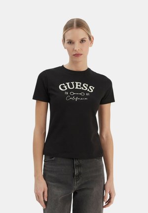 Donna con capelli biondi che indossa una maglietta nera GUESS e jeans grigio scuro, in piedi davanti a uno sfondo bianco semplice.