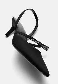 Svart slingback-högklackad sko med spetsig tå. Har satinliknande material, tunn ankelrem och en kristallsmyckad rosettdetalj.