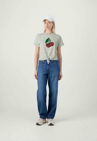 Striped t-shirt in beige and green with a sequin cherry design, knotted at the waist. Paired with blue jeans and light-colored sneakers.