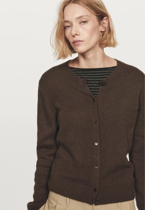 Brauner Strickcardigan mit runden Ausschnitt, gerippten Bündchen und Knöpfen vorne. Über einem gestreiften Shirt in Grüntönen getragen.