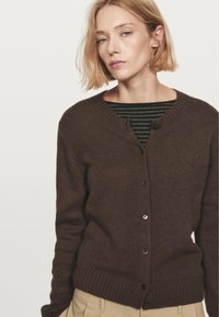 Brun strikket cardigan med rund hals, ribbede manchetter og knapper foran. Laget over en stribet skjorte med grønne nuancer.