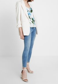 Blazer blanco sobre una blusa floral con acentos en azul y verde, combinada con jeans ajustados azul claro y sandalias de tacón blancas.