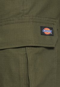 Olivenfarget stoff med en teksturert overflate, som har et Dickies-logo merke og synlige sømdetaljer på lommeområdet.