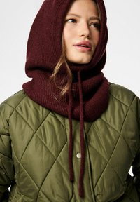 Grüne gesteppte Jacke mit einer burgunderfarbenen Strickkapuze. Die Kapuze hat eine gerippte Struktur und Kordelzüge; die Jacke verfügt über einen Druckknopfverschluss.