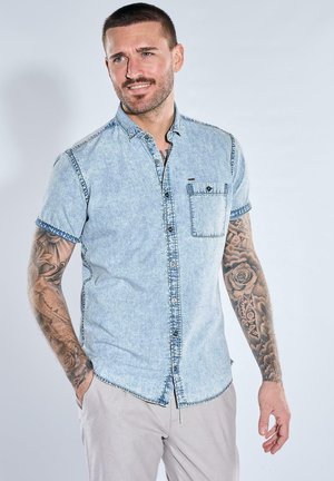 Mann mit kurzen Haaren und Tätowierungen auf beiden Armen, trägt ein helles Jeanshemd mit kurzen Ärmeln und beige Hosen, steht vor einem schlichten Hintergrund.