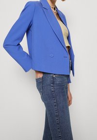 Veste bleu courte avec épaules structurées, dotée d'une fermeture à double bouton et d'une texture de tissu lisse, associée à un jean taille haute.
