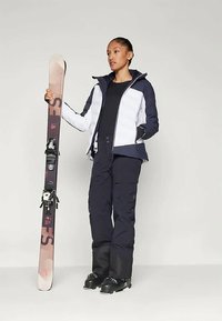 Lyžiarsky outfit pozostávajúci z bielej a námorníckej izolovanej bundy, čierneho trička a námorníckych nohavíc. Osoba drží vzorovanú lyžu so viazaním.