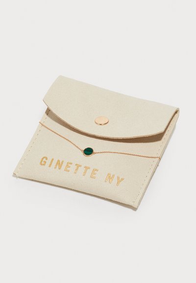 Beige keinonahka-pussi, jossa on kultainen napin sulku. Siinä on "GINETTE NY" präntätty kultaisena ja vihreä pyöreä korukivikoriste ketjussa.
