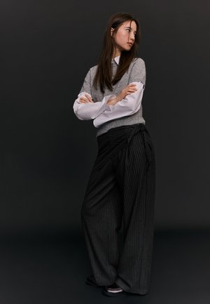 Frau steht mit verschränkten Armen, trägt einen grauen ärmellosen Pullover über einem weißen Hemd und weit geschnittene dunkle Nadelstreifenhosen vor schwarzem Hintergrund.