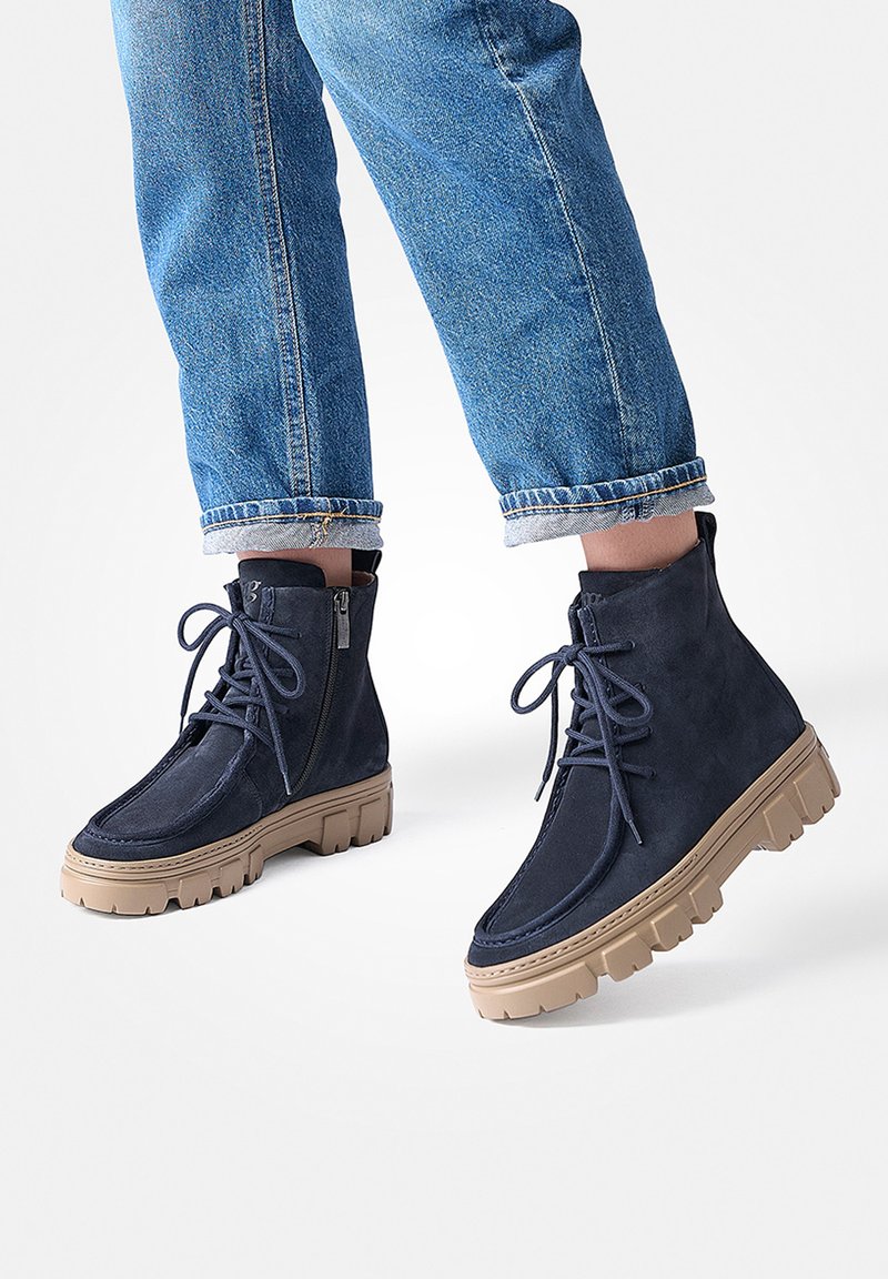 Des bottines en suède bleu marine avec une semelle épaisse beige, un laçage à l'avant et une fermeture éclair sur le côté pour un enfilage facile, associées à un jean bleu retroussé.
