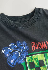 Gros plan d'un sweat-shirt noir à col rond montrant un texte coloré de style graffiti avec les mots "BOOM" en rouge et "CREEPER" en vert.