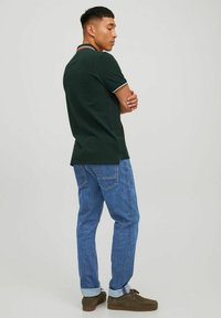 Maglietta polo verde scuro con colletto e rifiniture a contrasto, abbinata a jeans in denim azzurro chiaro. Il tessuto sembra morbido, con una vestibilità casual.