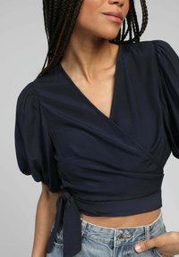 Femme avec des cheveux tressés portant un crop top en wrap bleu marine à manches bouffantes et un jean en denim bleu clair, la main reposant sur la hanche.