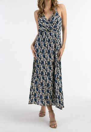 Femme portant une robe sans manches, longueur cheville, à imprimé serpent, avec un décolleté en V croisé, et des talons beiges à bride à la cheville, debout devant un fond blanc.